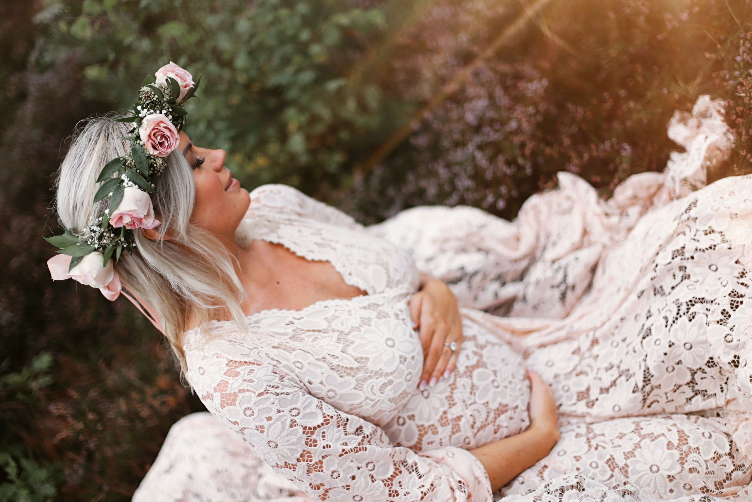 Artistic maternity portrait with white lace gown and rose crown in a garden setting.