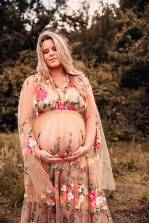 Maternity photo featuring floral dress against natural outdoor backdrop.
