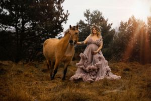 Ethereal maternity photo shoot with a brown horse and flowing mauve ruffled dress in a forest at sunset.