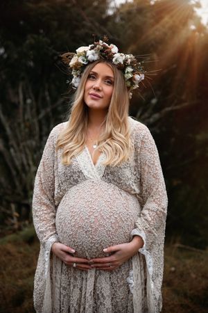 Maternity portrait featuring a delicate lace dress and floral crown with sunlight streaming through trees.