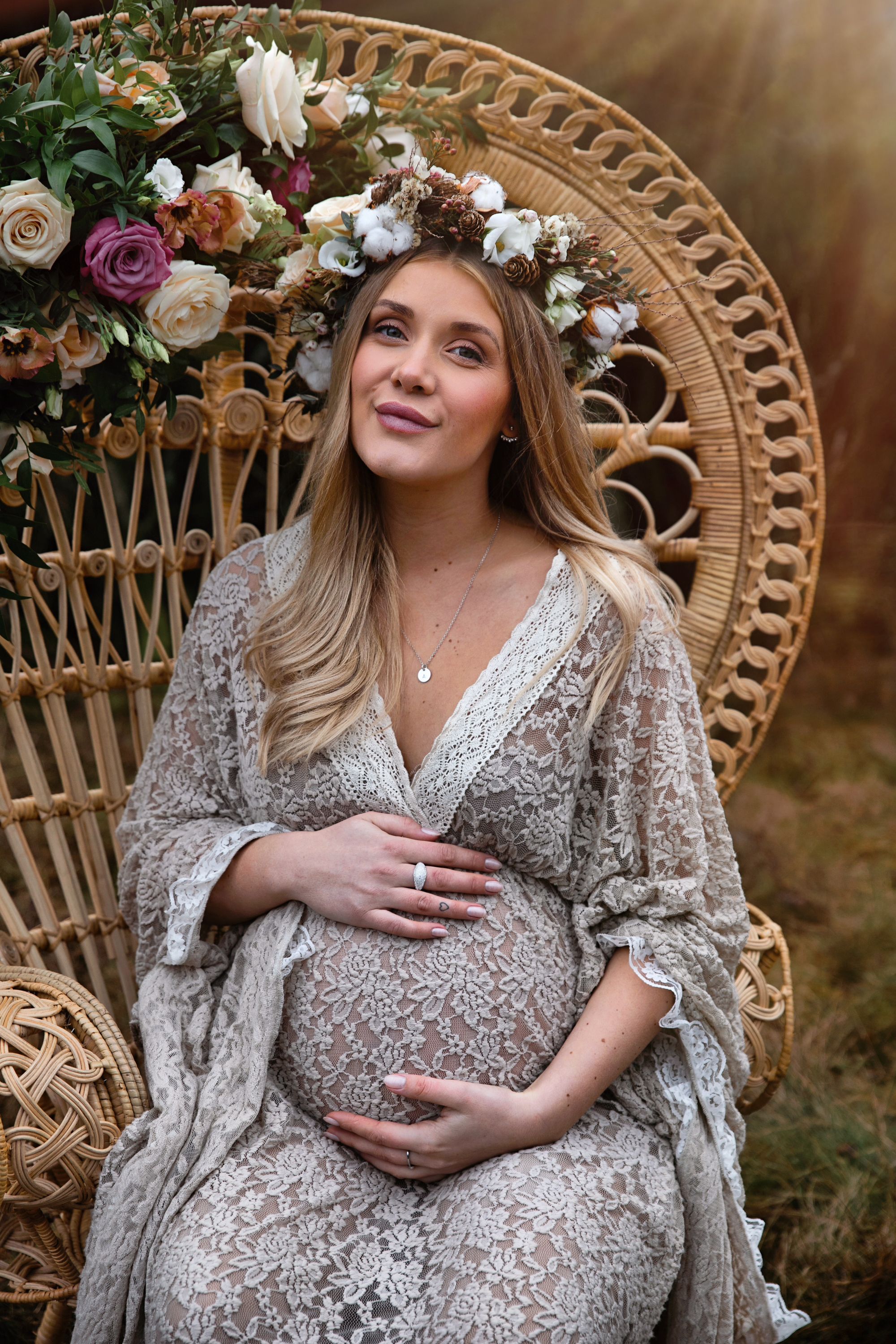 Joyful maternity portrait in a peacock chair wearing lace dress and floral crown.