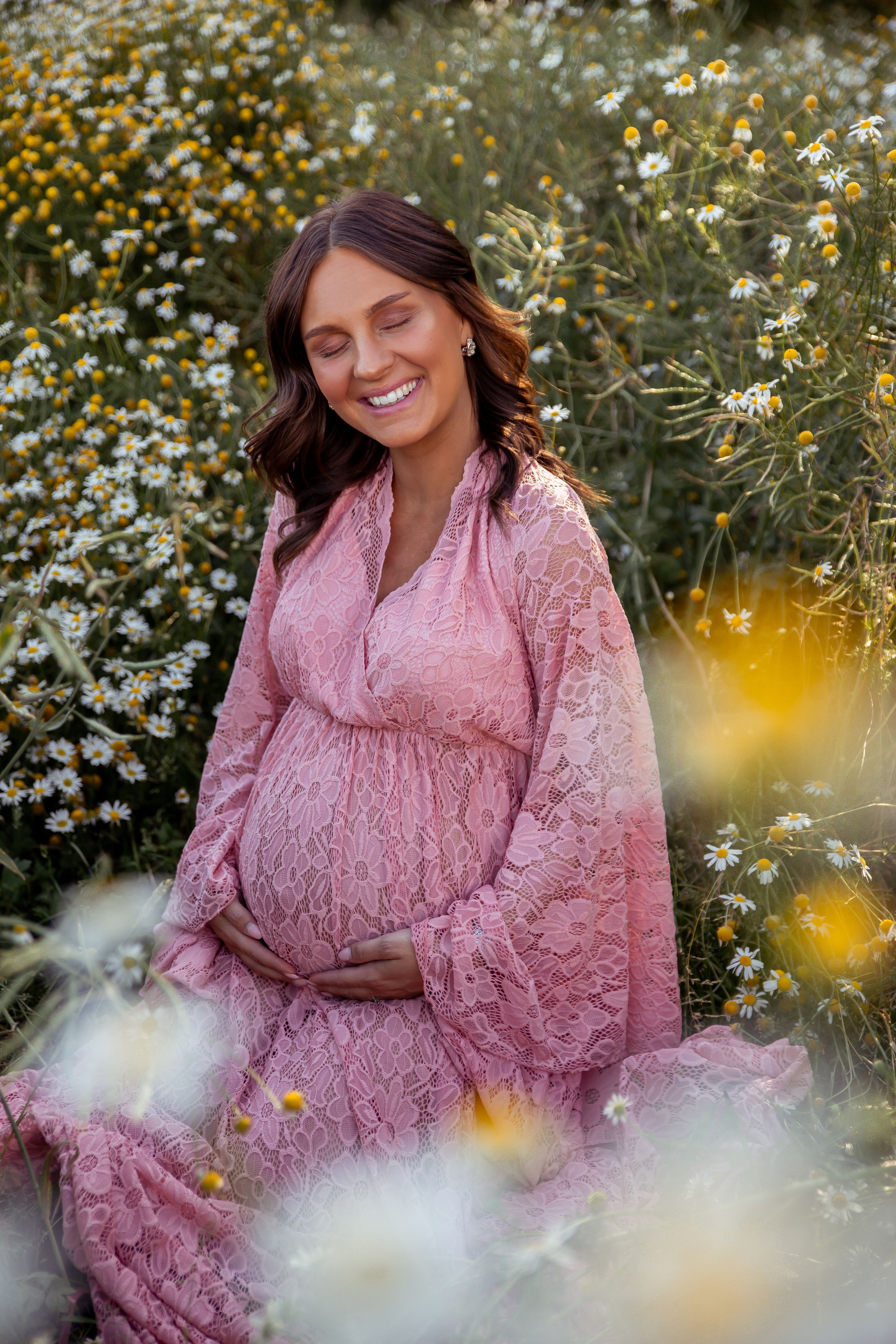 Pregnant woman in pink lace dress smiles while sitting in field of daisies.