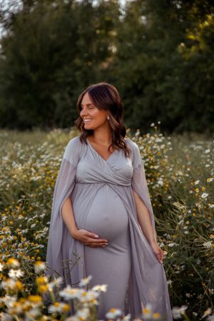 Maternity photo of woman in grey wrap dress standing among blooming wildflowers.