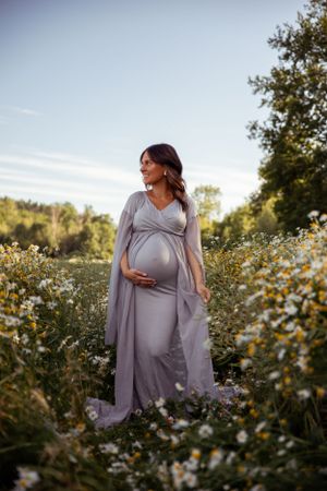 Elegant maternity photo of woman in grey dress standing among wildflowers at sunset.
