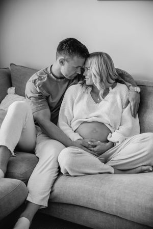 Intimate black and white maternity photo of couple sitting on couch with expectant mother showing bare pregnant belly.