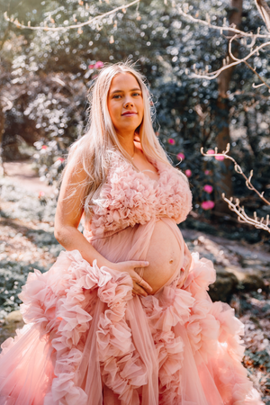 Maternity photo featuring expectant mother in flowing pink ruffled gown amid blooming spring branches.