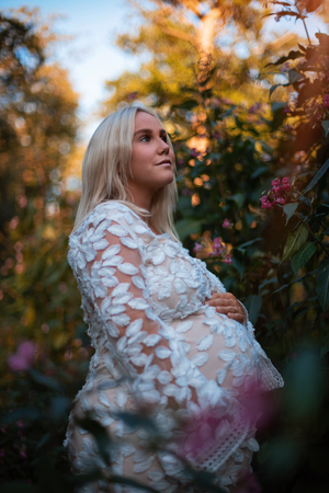 Maternity portrait of woman in white lace dress among autumn foliage at sunset.