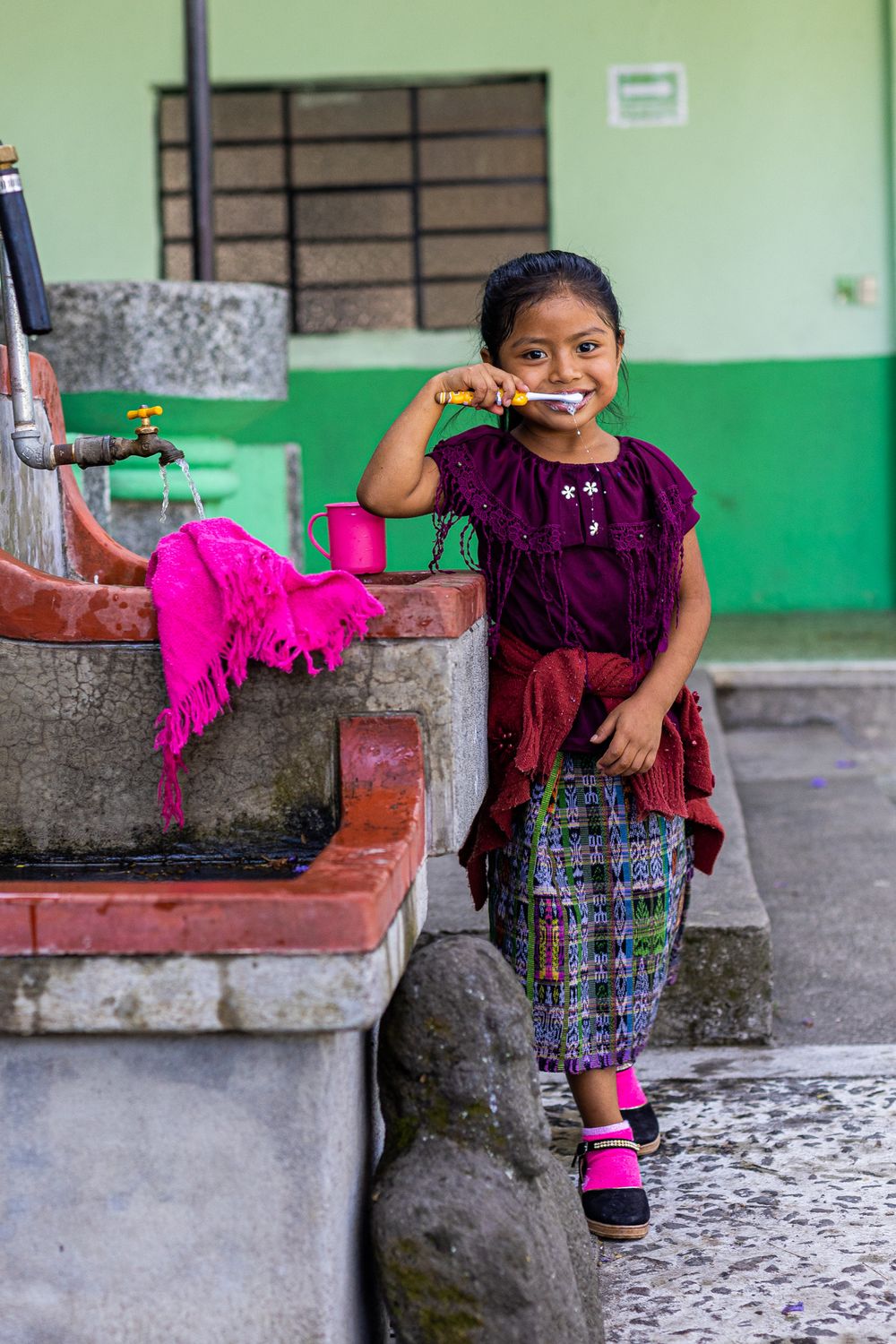 Young child in traditional Guatemalan dress smiling while brushing teeth at an outdoor sink with bright pink fabrics nearby.