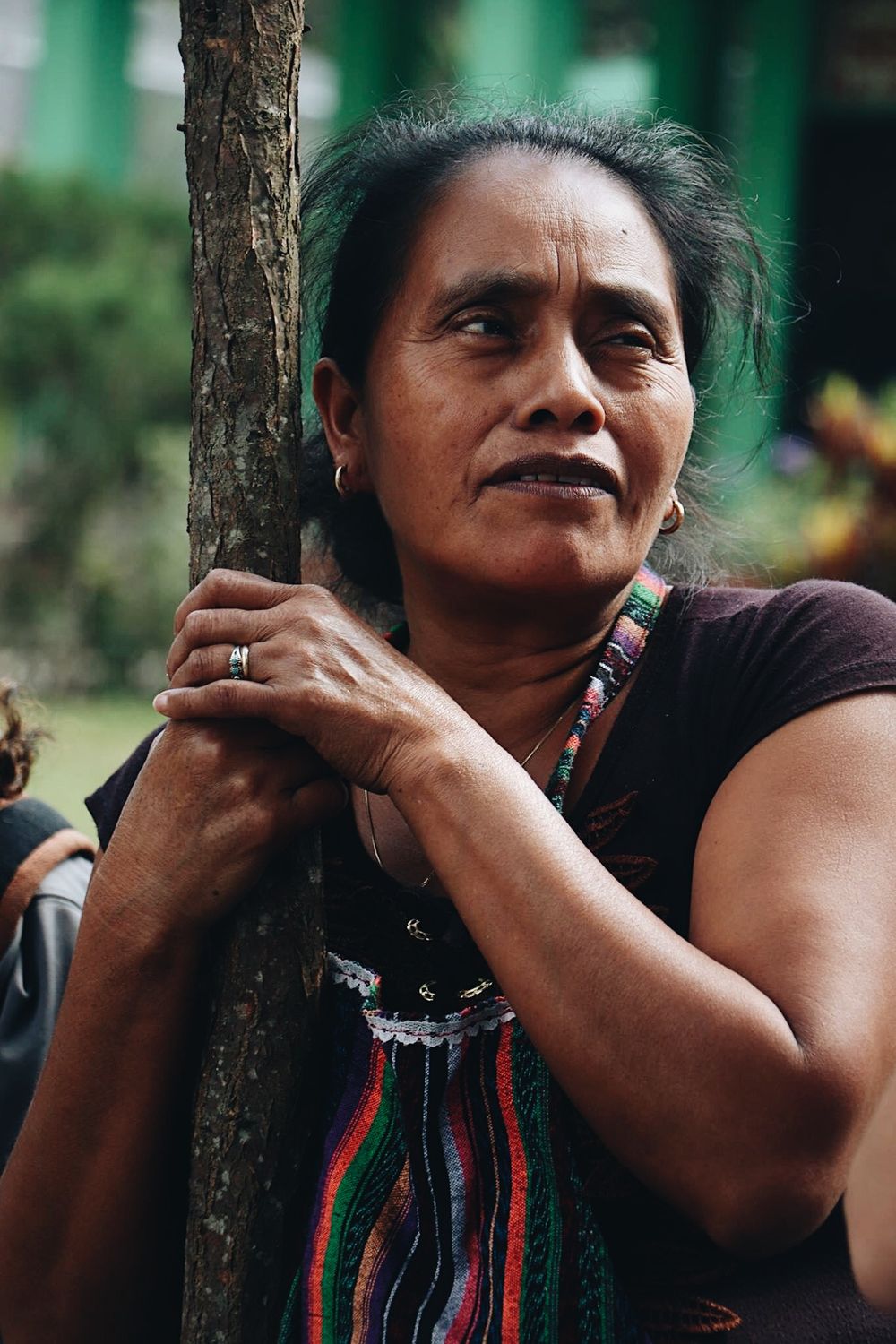 Woman in traditional Guatemalan dress leans against tree trunk while wearing colorful woven textiles.