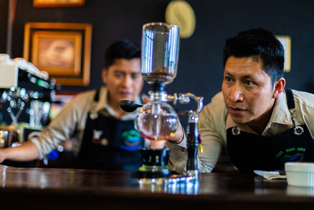 Two coffee professionals carefully examining a siphon brewing setup in a dimly lit cafe environment.