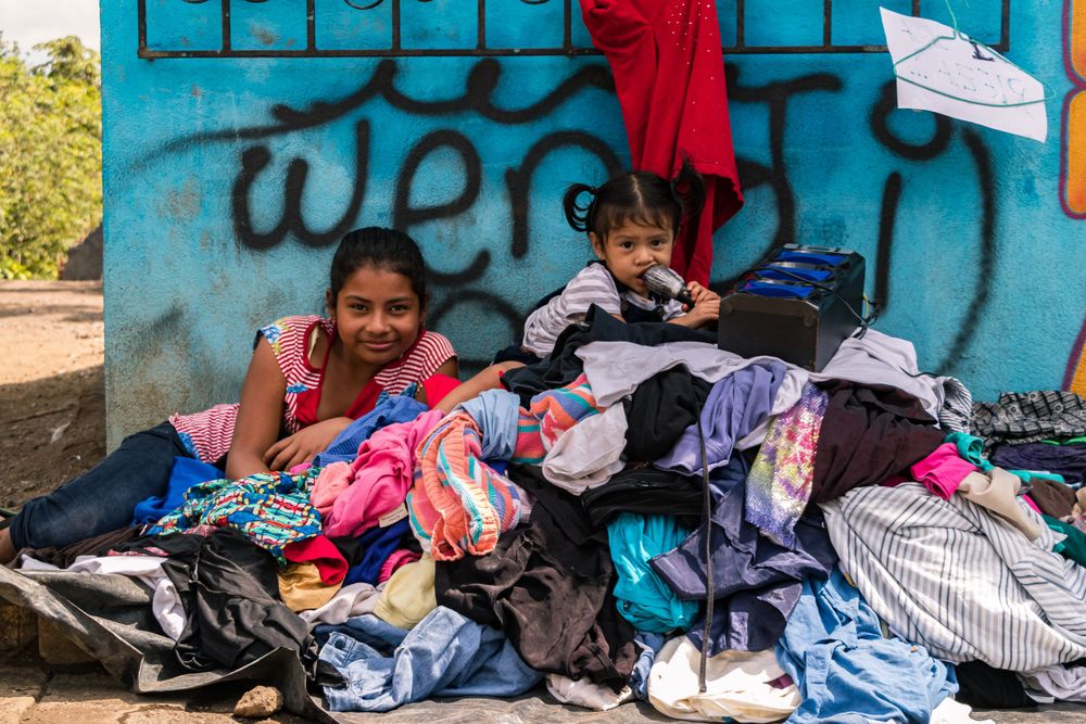 Piles of colorful laundry lay sorted against a blue graffiti wall outdoors.