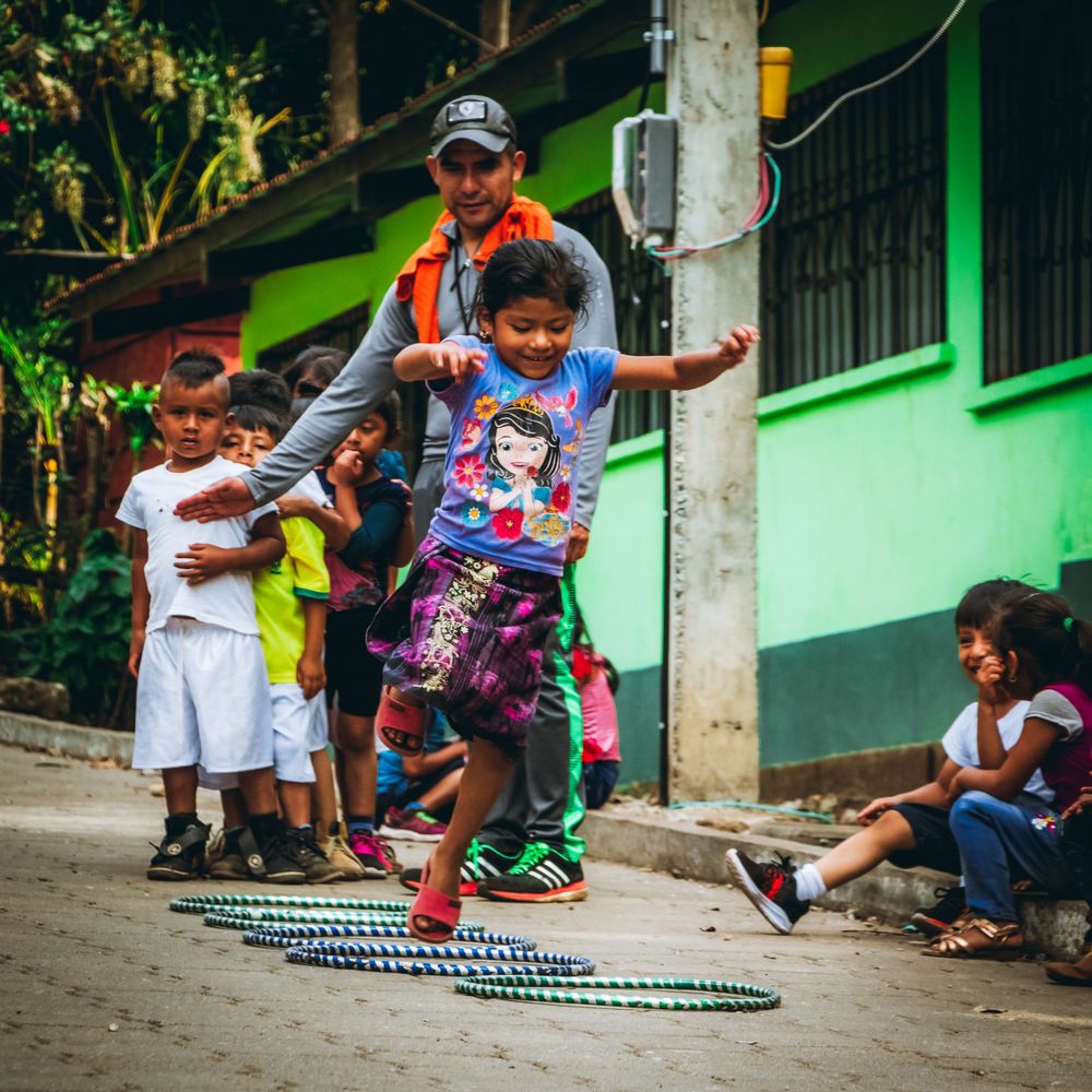 Children playing and jumping on hopscotch patterns on a street with green buildings in the background.