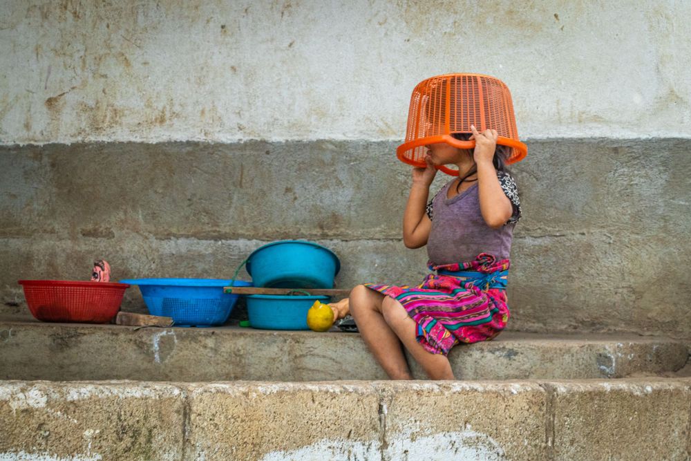 Child sitting on concrete steps with plastic containers and orange bucket over head.