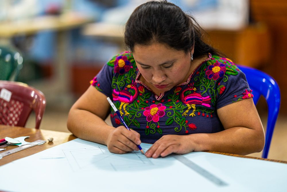 Person in colorful traditional embroidered dress writing on paper at a blue table.