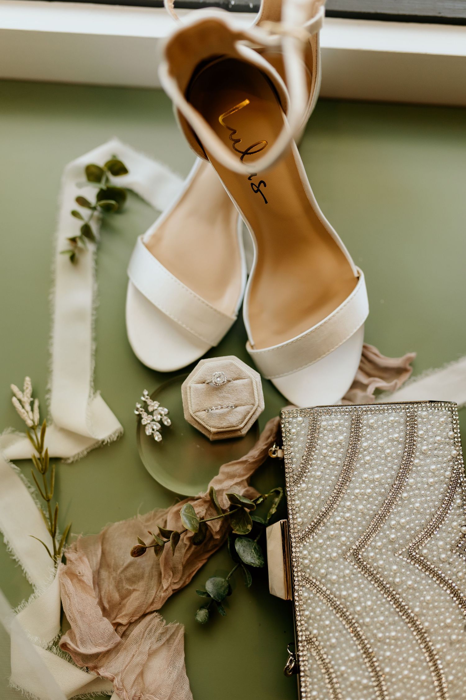 Wedding accessories including white heels, jewelry box and beaded clutch on green surface.