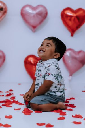 Child in floral print shirt lying on floor with scattered red hearts and balloons.