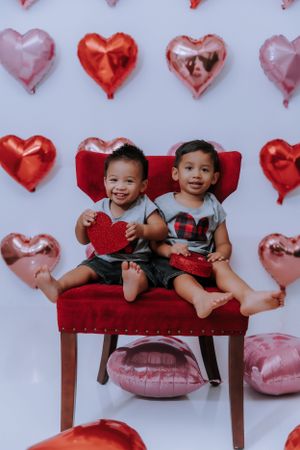 Two young children pose playfully on a red cushion against a backdrop of heart-shaped balloons.