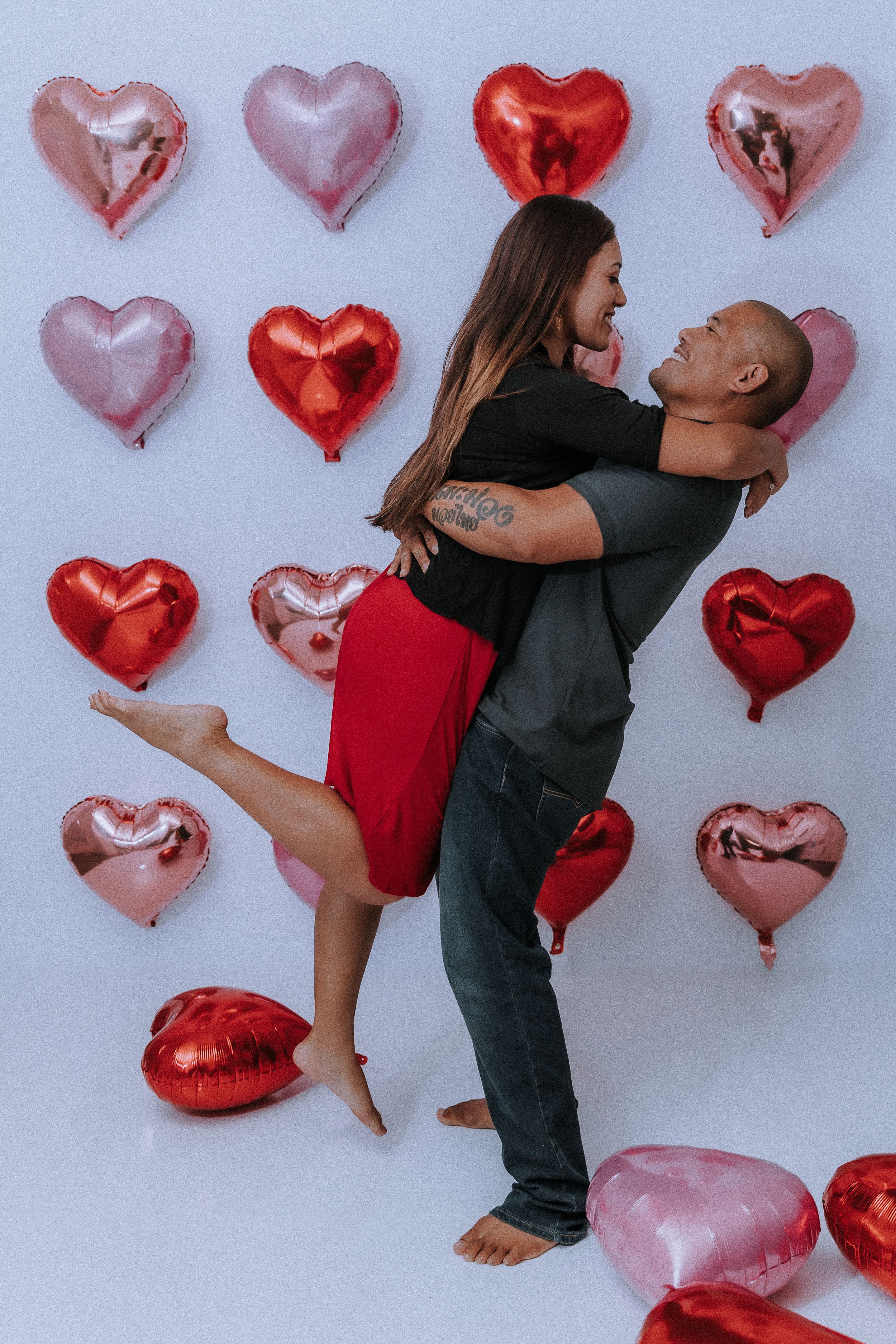 A couple poses playfully against a wall decorated with pink and red heart-shaped balloons.