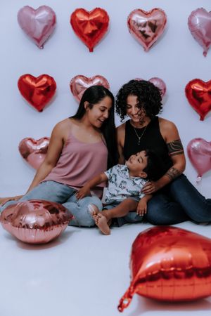 A couple lounges together on red cushions surrounded by heart-shaped balloons.