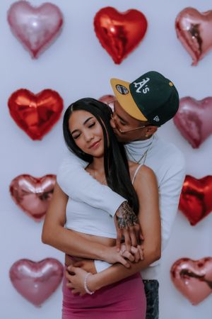 Couple embracing against wall decorated with red and pink heart balloons.