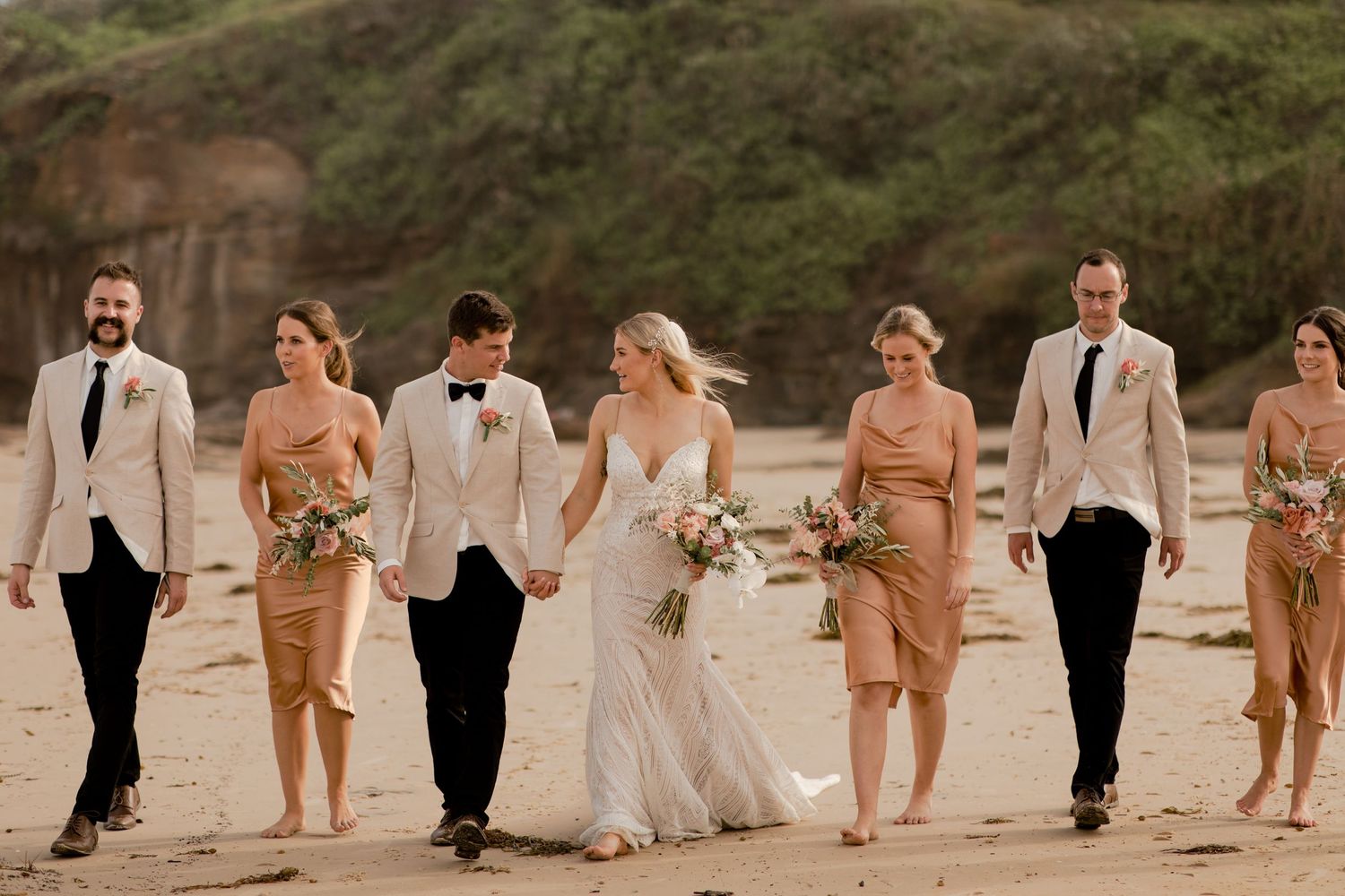 Wedding party walking together on a beach with bridesmaids in peach dresses and groomsmen in light jackets.