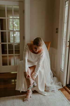 Bride in white dress fastening her shoe while sitting in sunlit room.