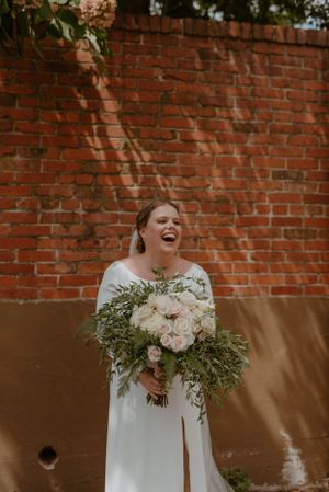 Joyful bride in white dress laughing against brick wall with bouquet.