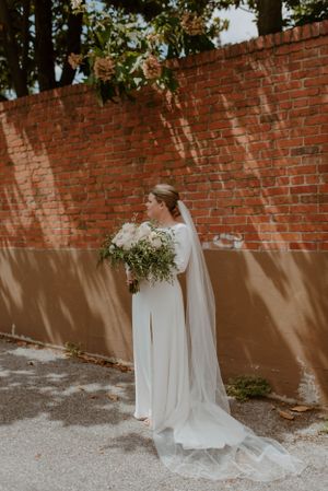 Elegant bride in flowing white dress and veil holds natural bouquet against rustic brick wall in natural lighting.