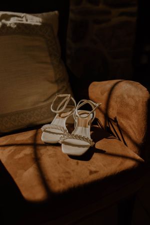 Elegant white bridal sandals with delicate straps catching sunlight on a brown textured surface.