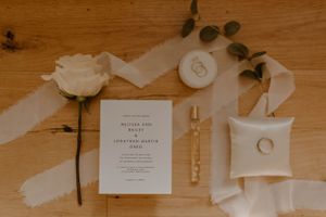 Wedding invitation flatlay with white rose, rings, and delicate ribbon on wooden surface.