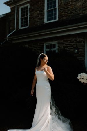 Elegant bride in fitted white gown and flowing veil poses against stone house backdrop.