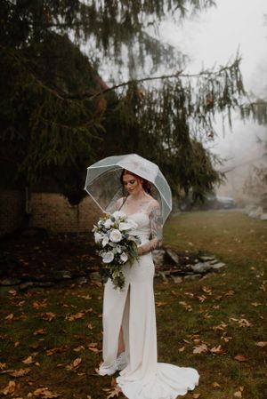 Bride holding clear umbrella and white bouquet poses in misty fall landscape.