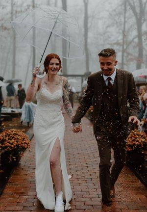 Joyful wedding couple walking through rain on brick path with clear umbrella and fall decorations.