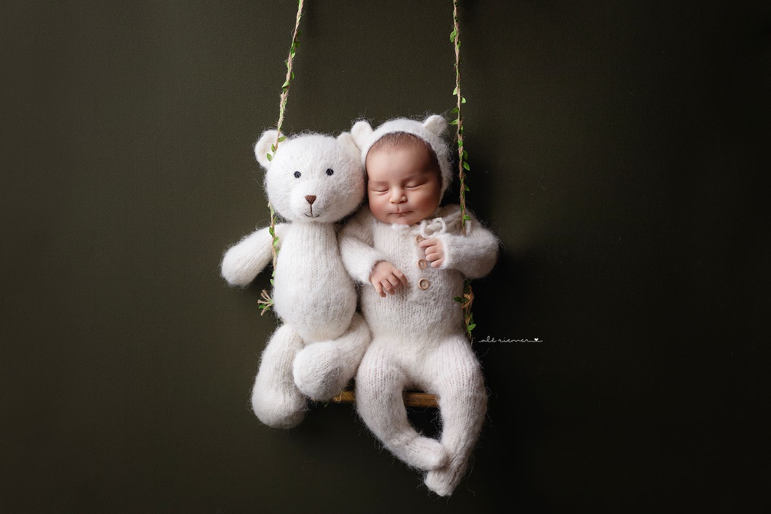 Creative newborn portrait with baby and teddy bear sitting together on swing against dark background.
