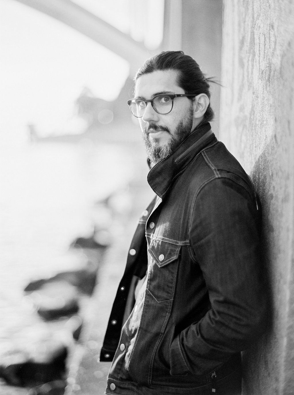 Black and white portrait of a bearded man in denim jacket leaning against a wall.