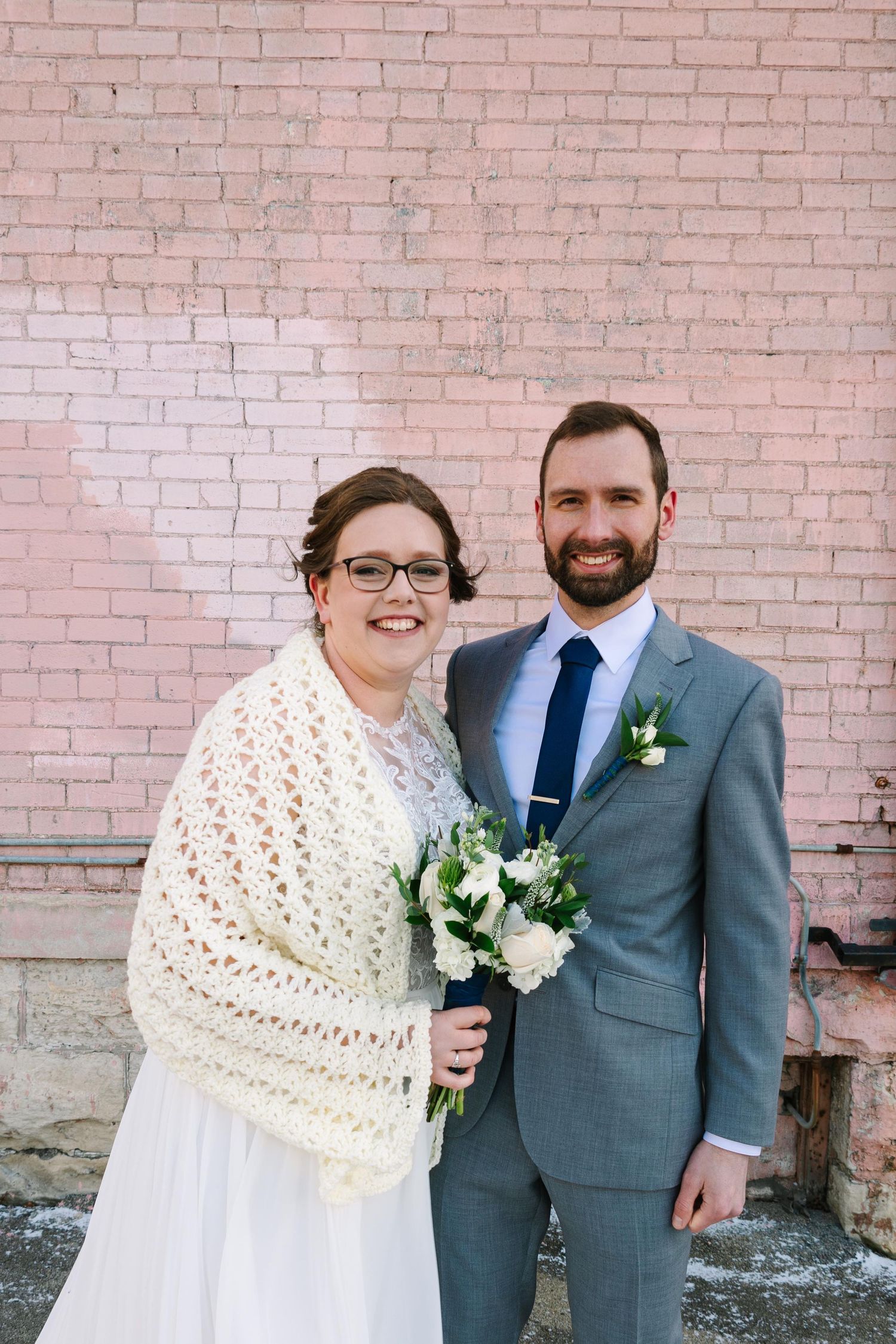 A winter wedding couple poses against a pink brick wall with a white knit shawl and floral bouquet.