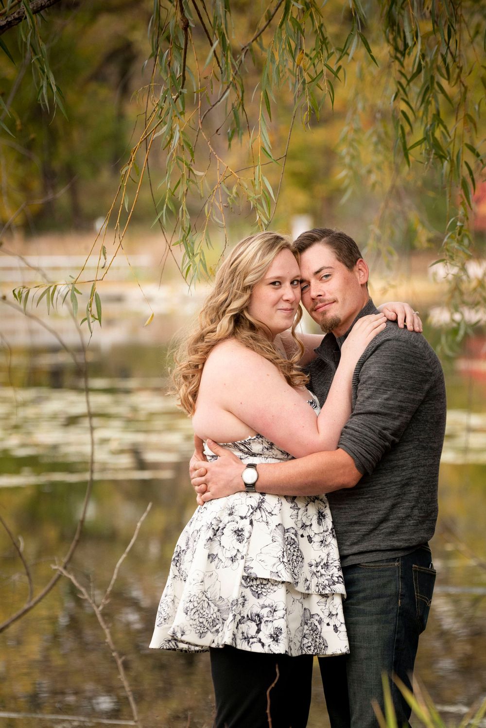 Couple embracing by pond with willow trees and autumn colors in background.