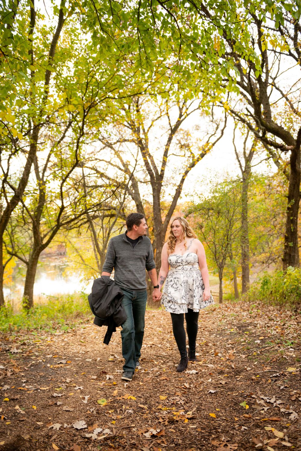 A couple strolls along a leaf-covered path beneath autumn trees.
