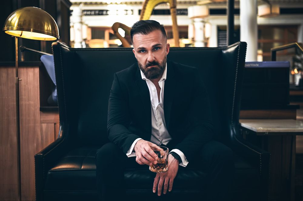 Bearded man in black suit holding whiskey glass seated in leather chair at upscale bar.
