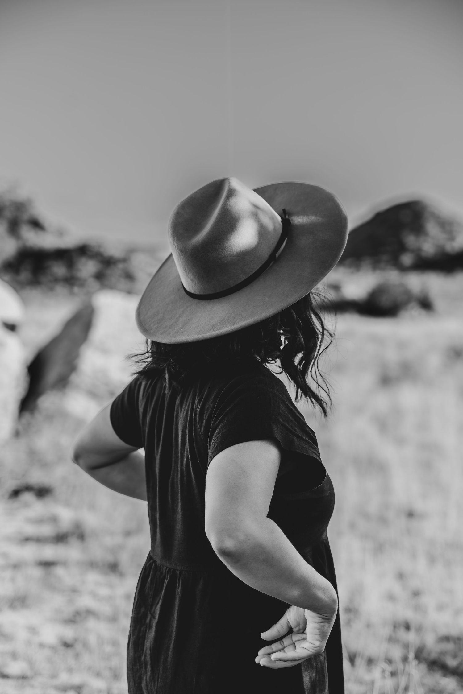 A person in black dress and wide brim hat stands in black and white desert landscape.