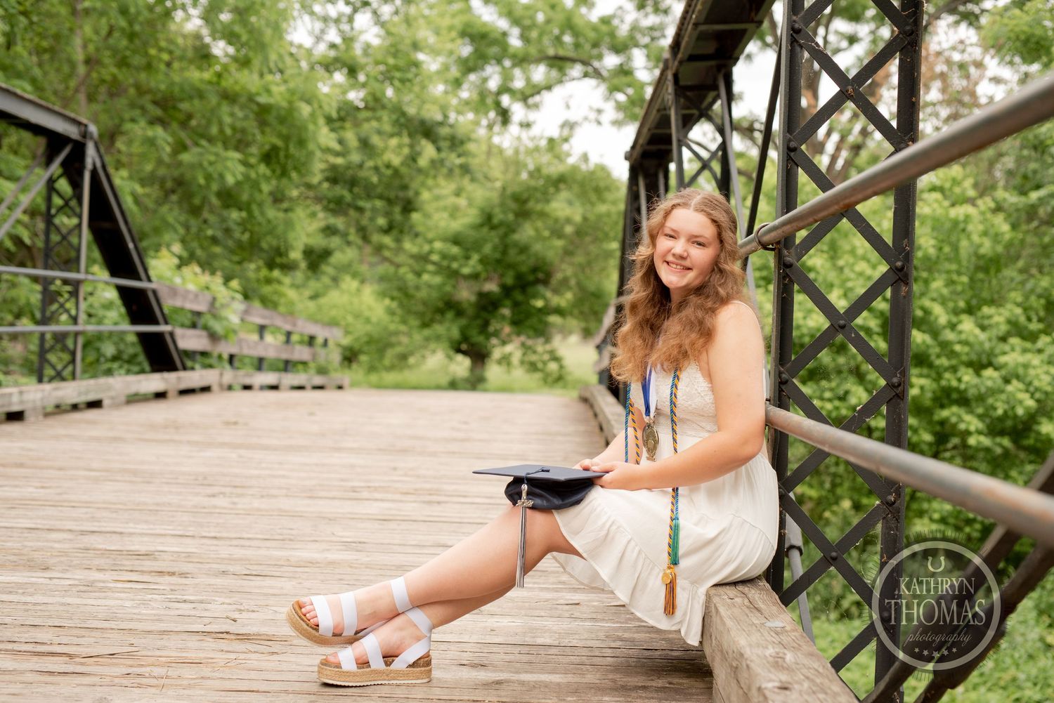 Graduate in white dress sits casually on rustic wooden bridge railing during photo session.