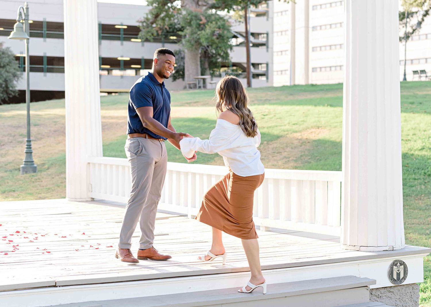 A couple stands together on a white gazebo porch with a parking structure visible in the background.