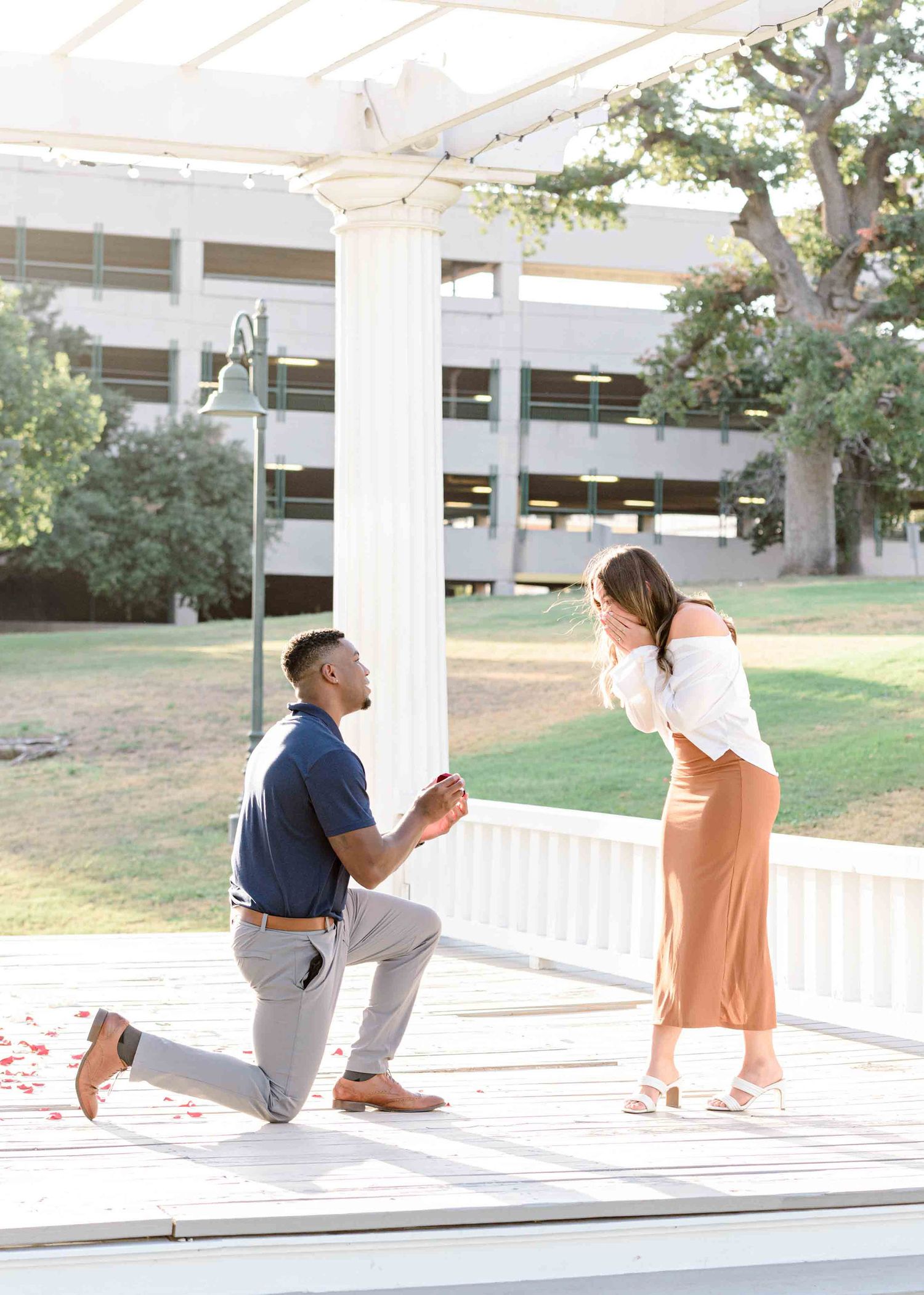 A proposal moment captured under a white gazebo with a parking structure in the background.