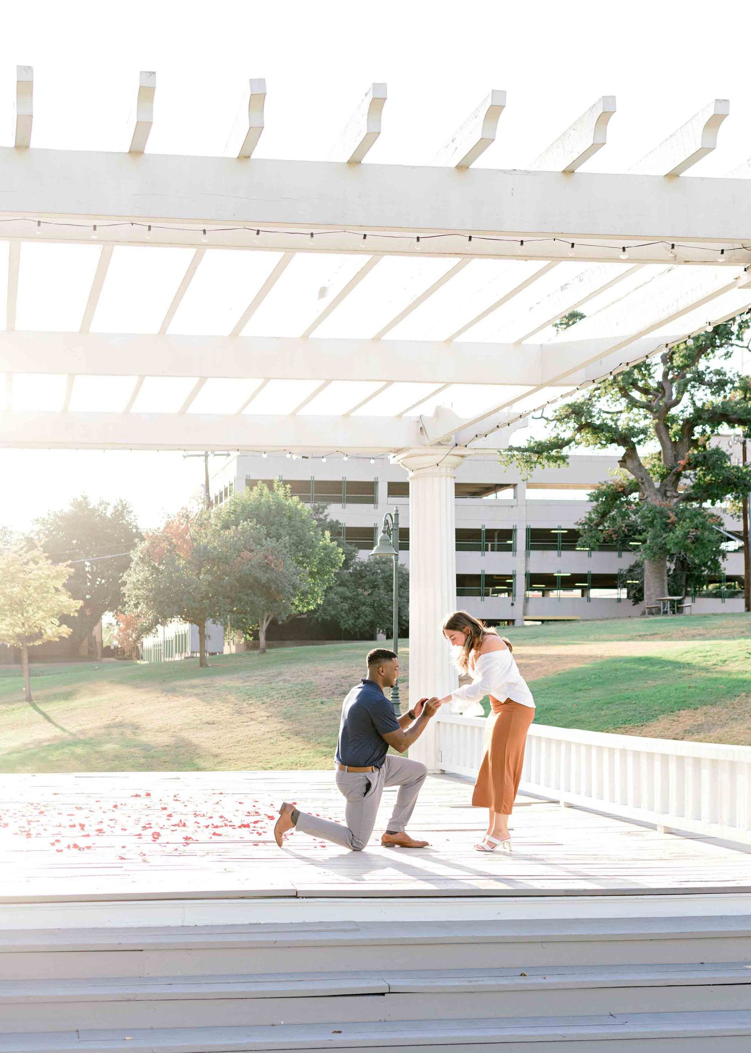 A sunset proposal scene under a white pergola with a parking structure and trees in the background.
