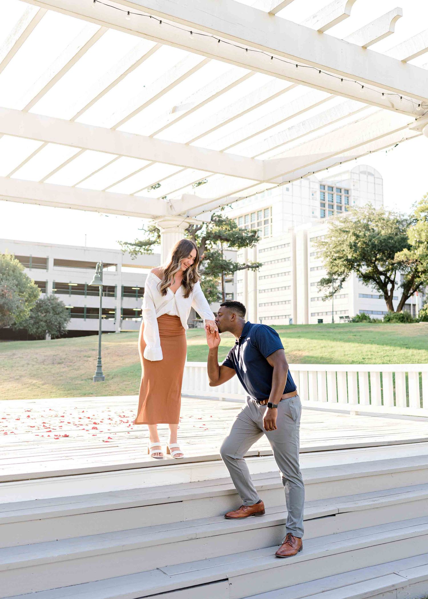 A romantic proposal scene under a white pergola with urban architecture in the background.