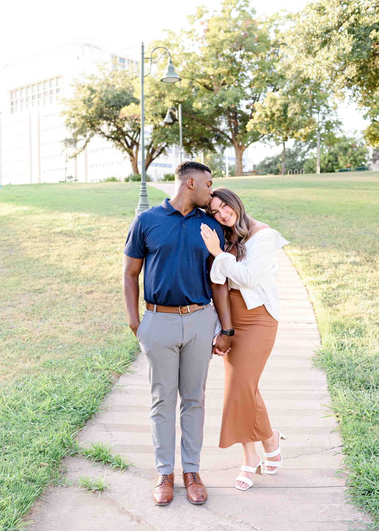 A couple sharing an intimate moment while standing on a pathway with trees in background.
