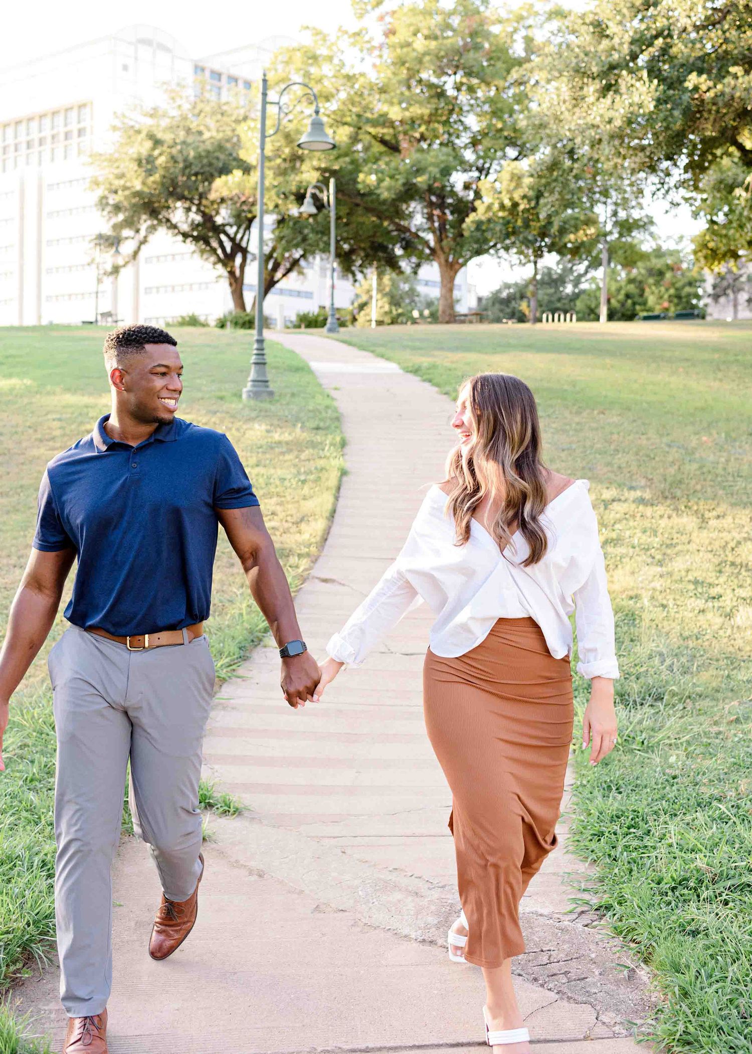 A couple holding hands while walking along a scenic pathway surrounded by grass and trees.