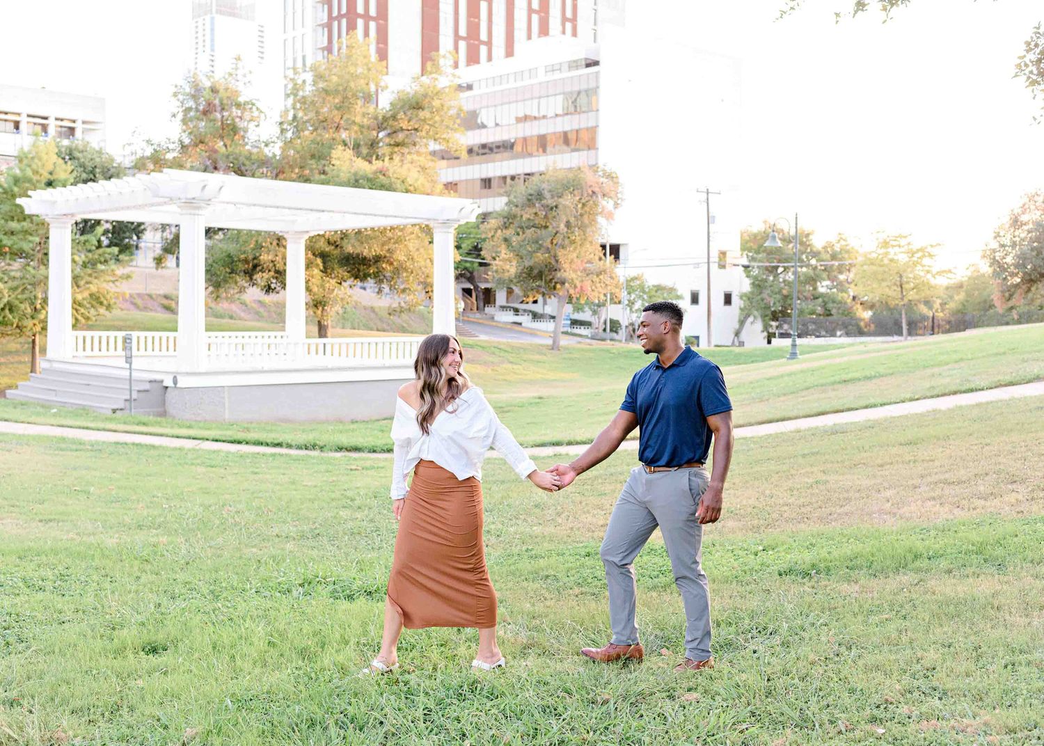 Couple holds hands and dances together in front of a white gazebo in a park.