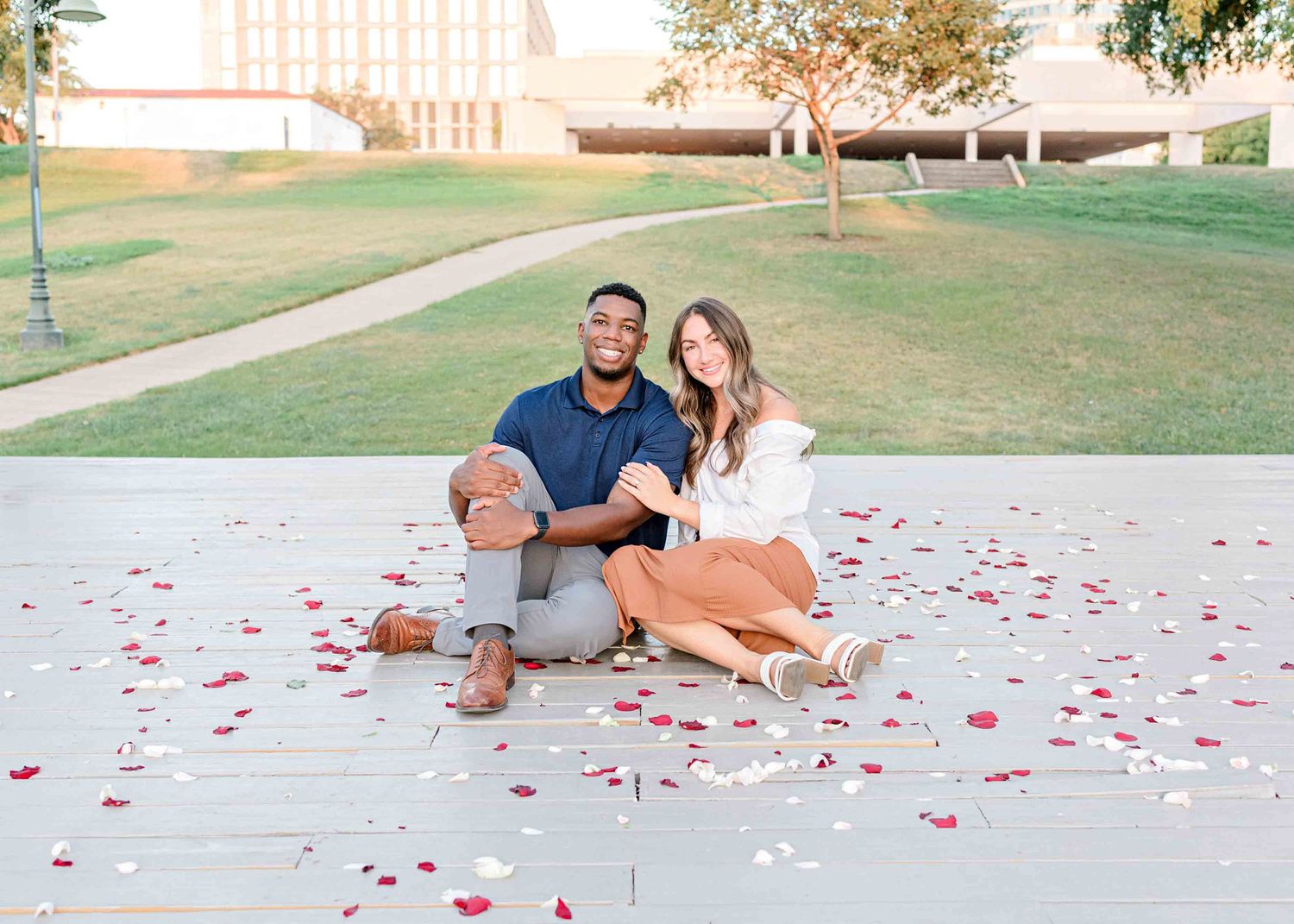 A couple sitting on ground surrounded by scattered rose petals in an outdoor setting.