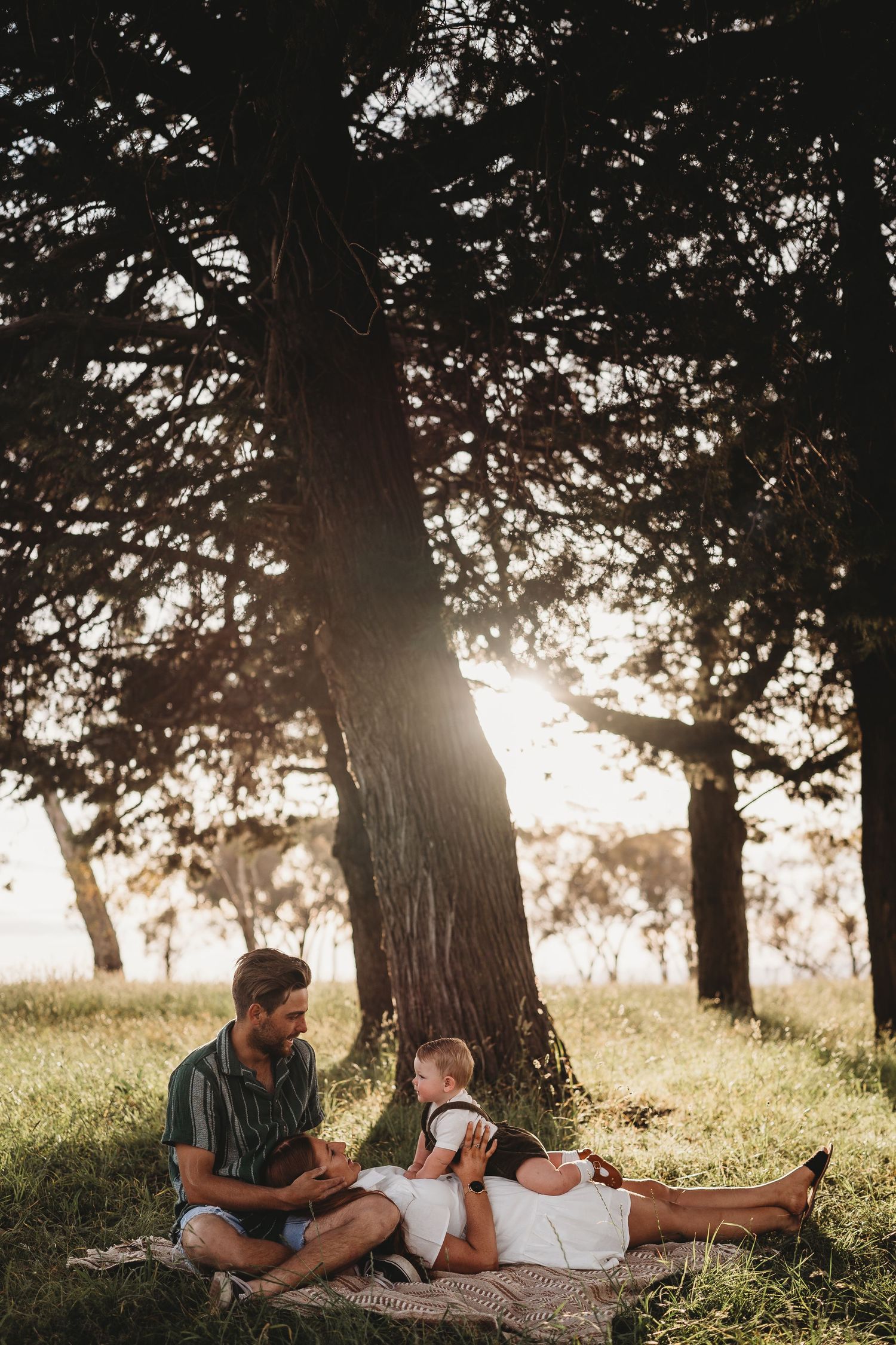 Family relaxing on a blanket under tall trees in golden sunset light in a park.